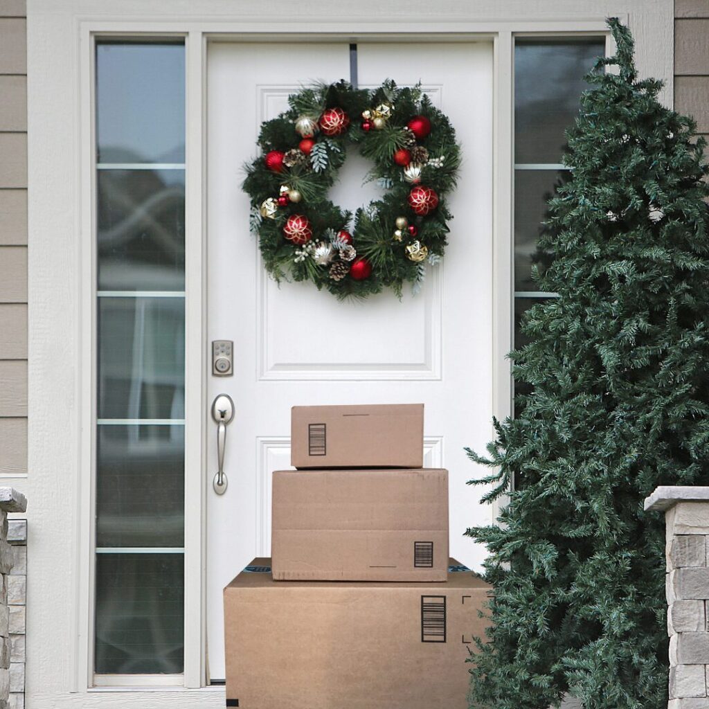 Front door decorated with a Christmas wreath, surrounded by stacked packages and a pine tree.