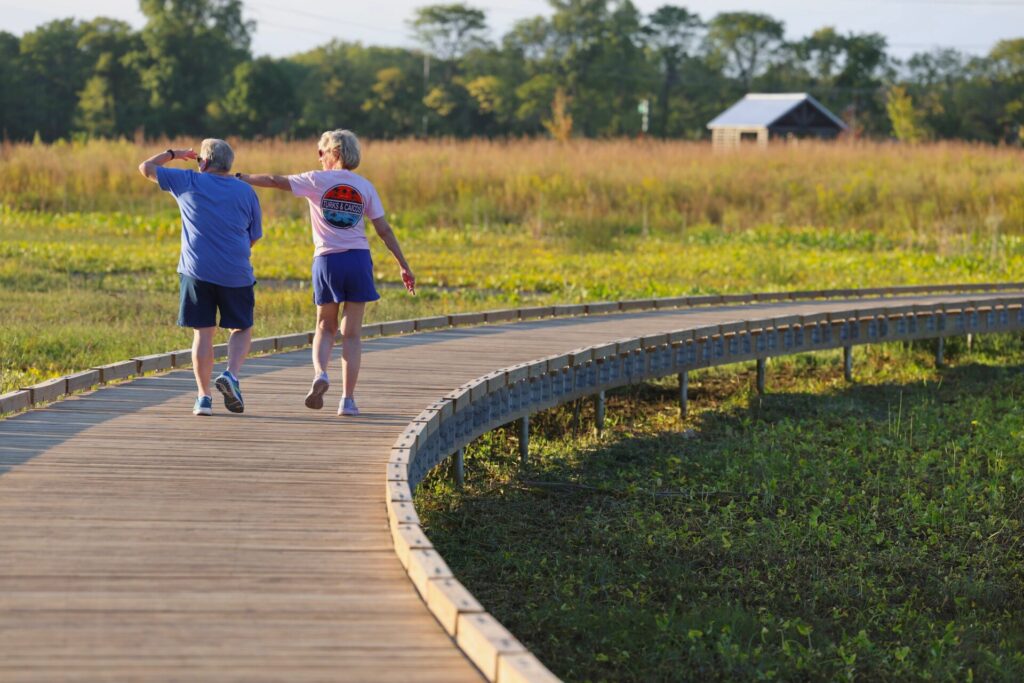 Two people walking on a curved wooden boardwalk in a grassy field on a sunny day.