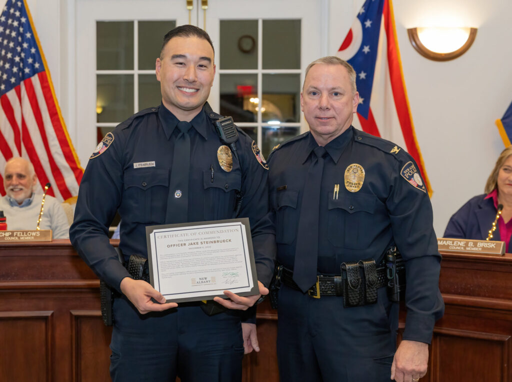 A police officer smiling and holding up a certificate of commendation with another officer by his side