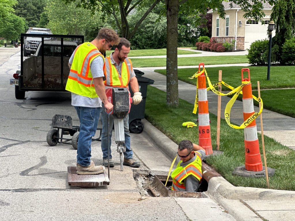 Three city workers fixing a sewer grate. One is holding a jackhammer, one is in the hole below street level, and one is standing on the old grate.