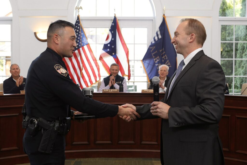 A police officer and a man in a suit shaking hands in a courtroom setting with people clapping in the background and several American flags displayed.