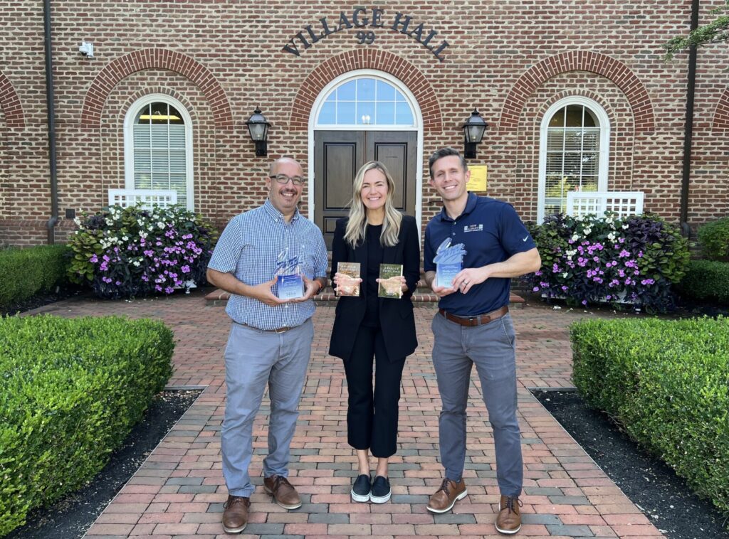 Three people standing in front of a brick building labeled "Village Hall," each holding awards and smiling.