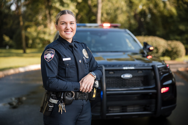 Police officer smiling and standing in front of a patrol car with lights on.