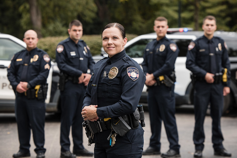 A group of police officers in uniform standing in front of patrol vehicles, with a female officer in the foreground smiling confidently.