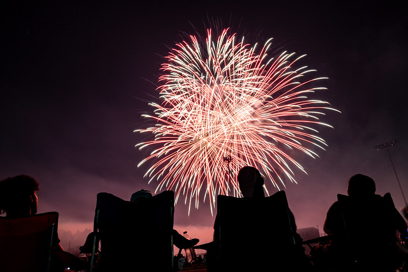 Silhouettes of people sitting on folding chairs watching a large display of red and white fireworks at night.