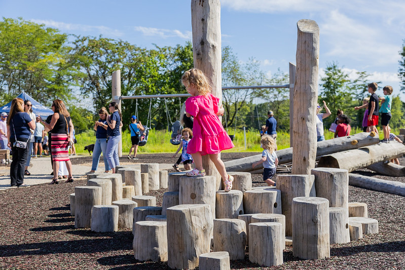 Children playing on wooden climbing structures at a playground, with adults and other kids in the background.