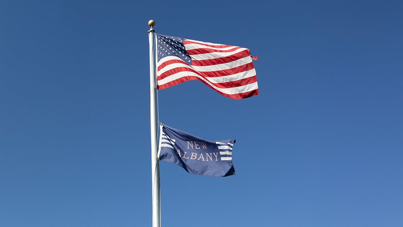 Two flags flying on a pole, with the American flag on top and a flag reading "New Albany" below, against a clear blue sky.