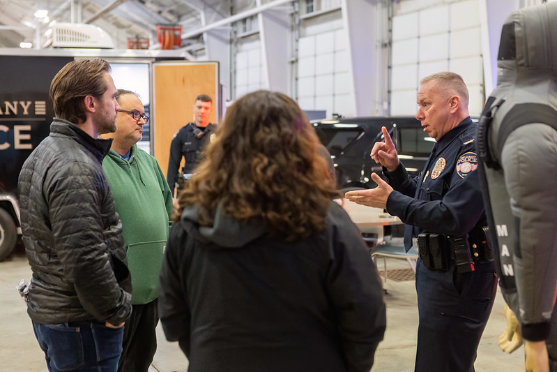 Police officer talks to a group of people inside a garage or workshop setting, with a police vehicle and equipment trailer in the background.