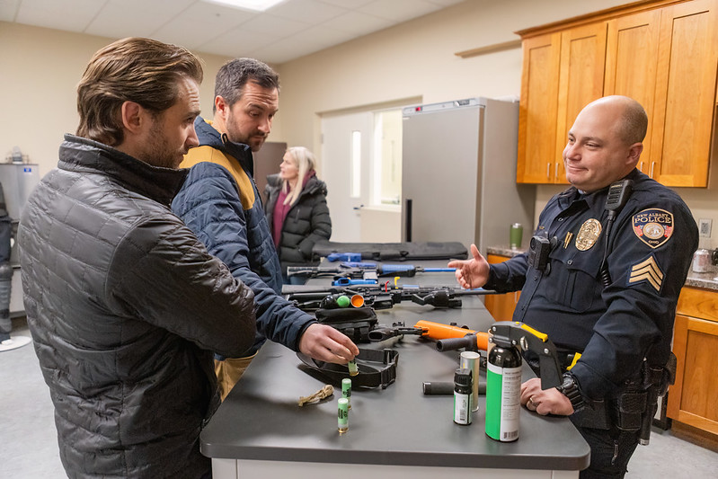 Three men, two in civilian jackets and one in a police uniform, are examining firearms and equipment on a table in a room with wooden cabinets and a refrigerator. A woman in a coat stands in the background.