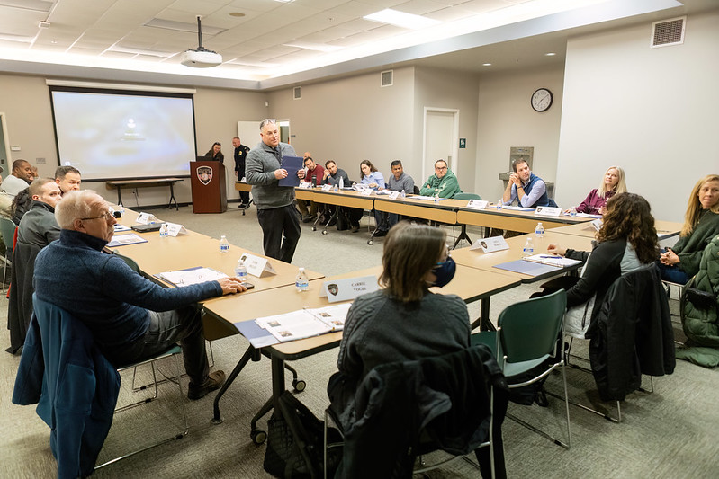 A group of people seated around tables in a conference room, participating in a meeting or workshop with one person standing and speaking.