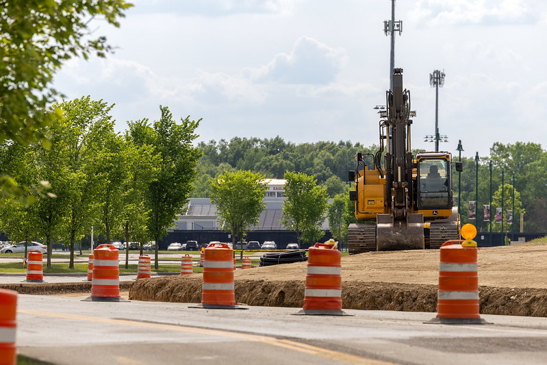 Construction site with an excavator and orange safety barrels, surrounded by trees.