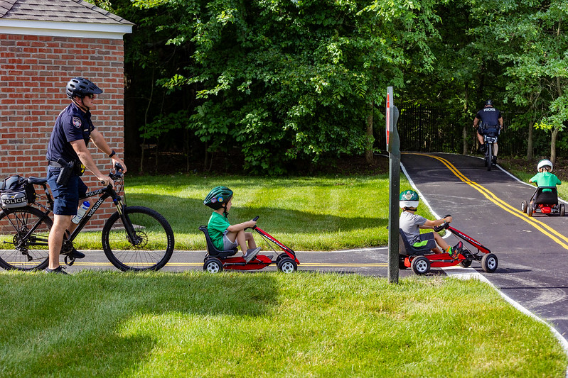 Police officers on bicycles interact with children riding pedal go-karts on a path in a green, wooded area.