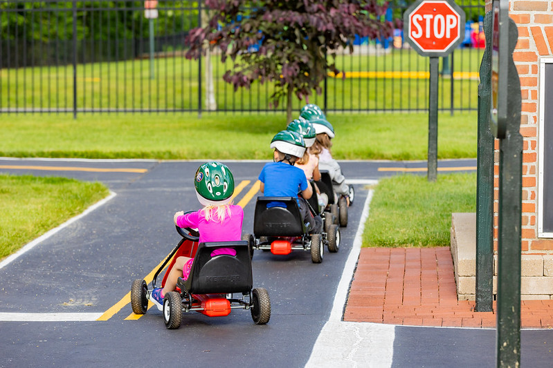 Children wearing helmets riding pedal go-karts on a miniature road with a stop sign.