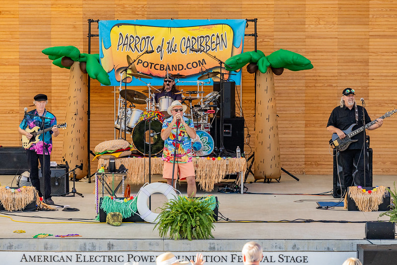 A band named "Parrots of the Caribbean" performing on stage with tropical decor, including palm trees and grass skirts. The band members are playing various instruments such as guitar, drums, and bass, with one member singing into a microphone.