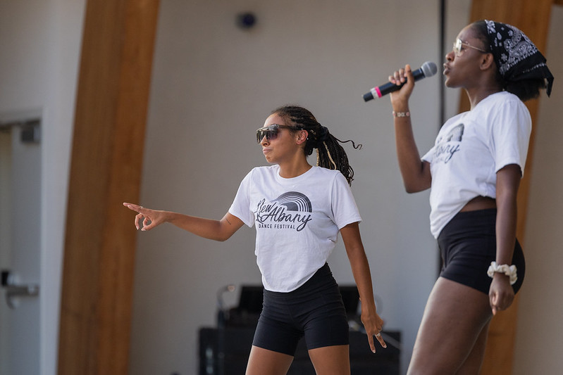 Two performers in "New Albany Dance Festival" shirts and black shorts, with one holding a microphone and the other pointing.