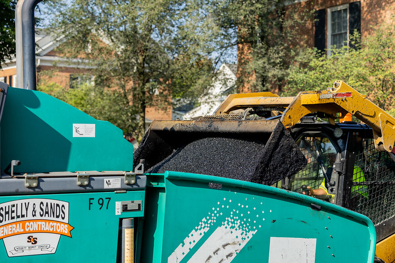 A construction vehicle pours asphalt into a green Shelly & Sands General Contractors machine.