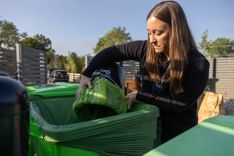 A woman disposing of a green bag into a green compost bin outdoors.