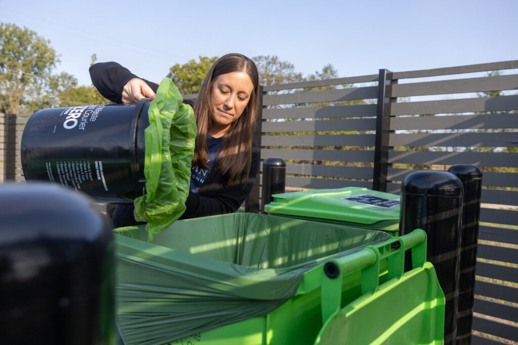 A woman empties a black compost bucket into a large green compost bin outdoors, with a wooden fence and trees in the background.