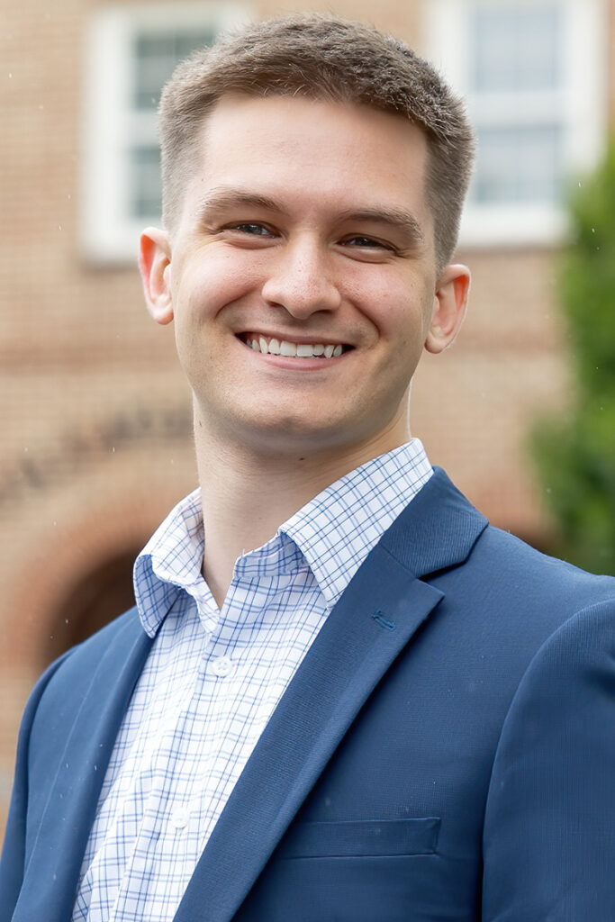 Smiling man in a blue blazer and checkered shirt standing outdoors.