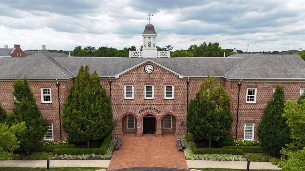 Village Hall building with a red brick exterior, arched entrance, clock above the main door, and a cupola on the roof, surrounded by green trees and a brick walkway.