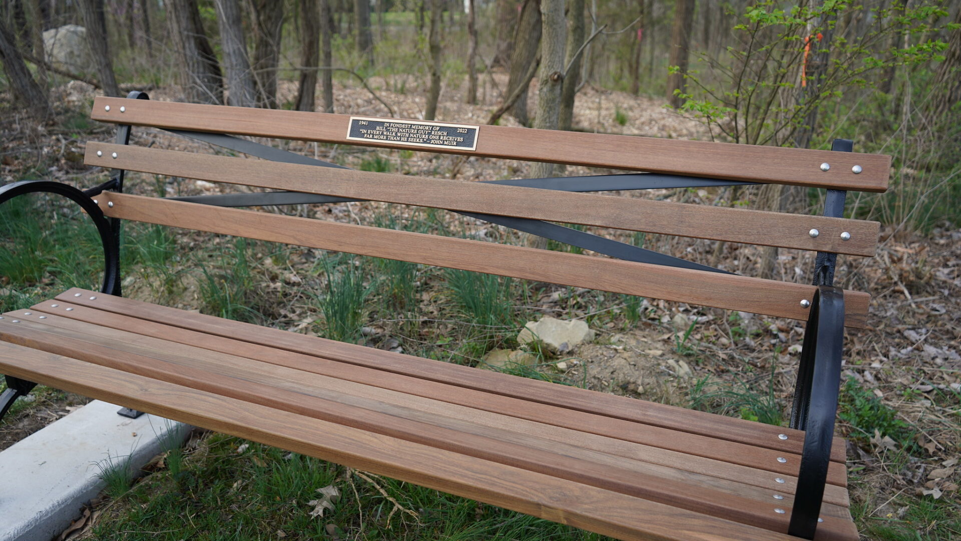 A wooden park bench with a memorial plaque, situated in a forested area.