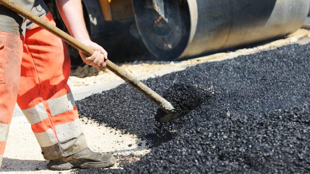 Worker spreading asphalt with a shovel next to a road roller.