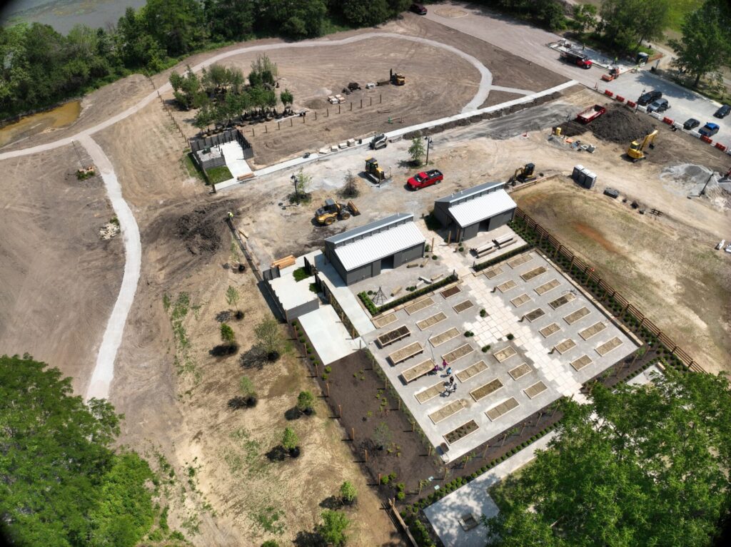 Aerial view of a construction site featuring several buildings, pathways, vehicles, and equipment. A garden area with rectangular plots is visible in the foreground. The site is surrounded by trees and natural greenery.