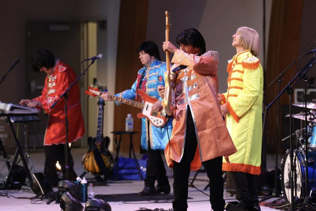 A band performing on stage dressed in colorful, retro-style costumes resembling Sgt. Pepper's Lonely Hearts Club Band outfits. One musician is playing a keyboard, another is on bass guitar, the third guitarist is adjusting his instrument, and the fourth member is standing beside them with a drum set in the background.