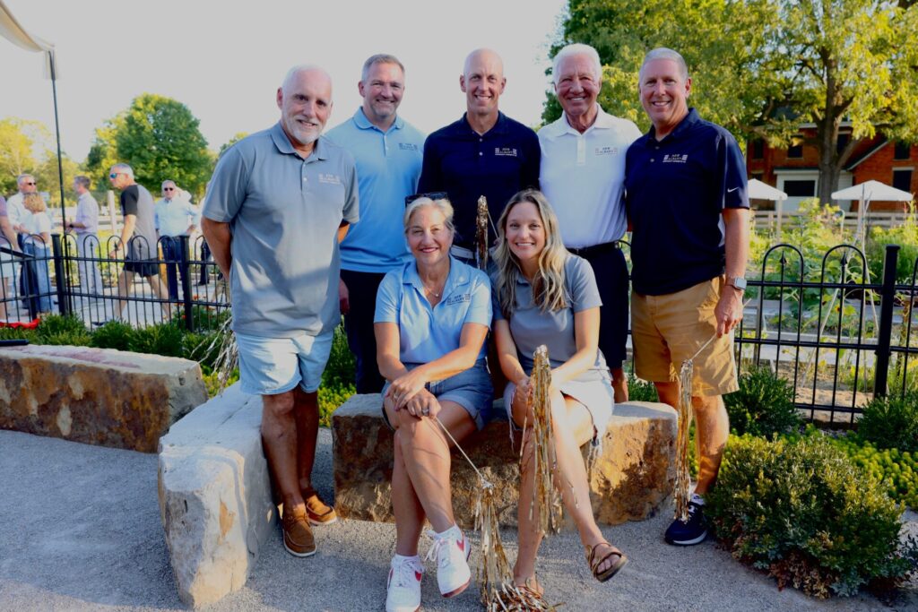 Group of seven people posing outdoors, with four men standing behind and one man and two women sitting on stone benches. They are casually dressed in polo shirts and shorts, and some are holding gold pom-poms. Trees and other people are visible in the background.