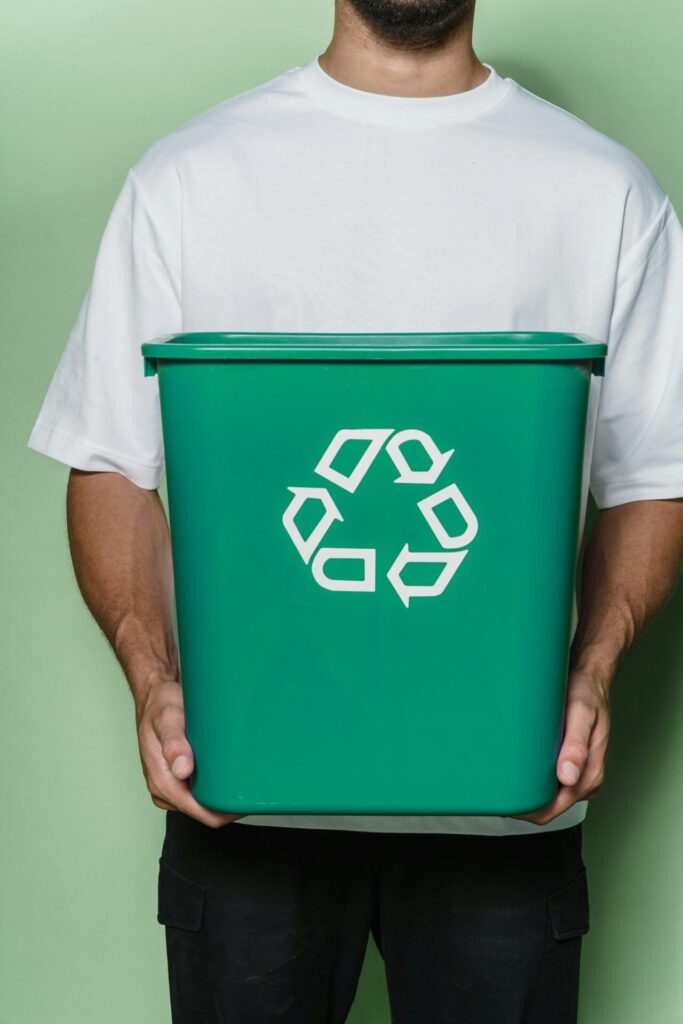 A person wearing a white t-shirt holding a green recycling bin against a light green background.