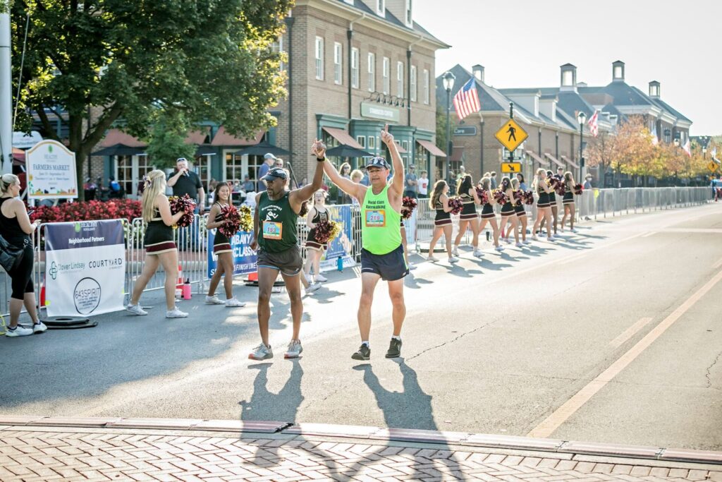 Two runners holding hands and celebrating as they cross the finish line of a race, with cheerleaders and spectators in the background.