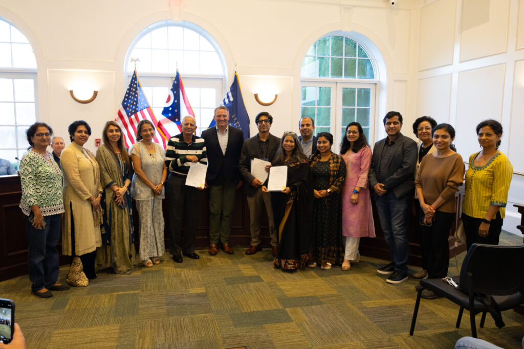 A group of people is standing indoors, posing for a photo in front of American flags and a window. Some individuals are holding certificates.