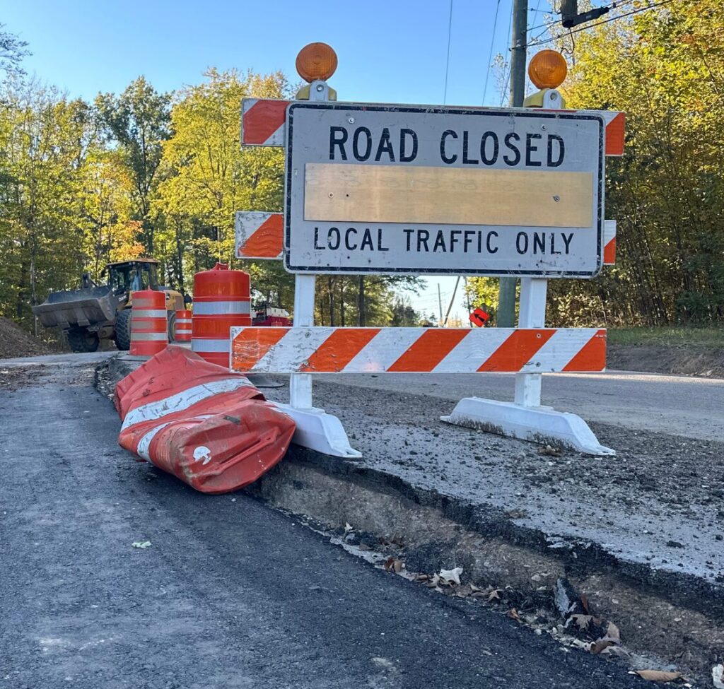 Roadwork scene with a "Road Closed" sign, orange barrels, construction equipment, and piles of dirt surrounded by trees.