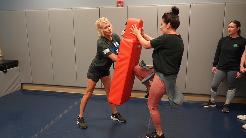 A woman practices a knee strike on a red pad held by another woman in a self-defense class.