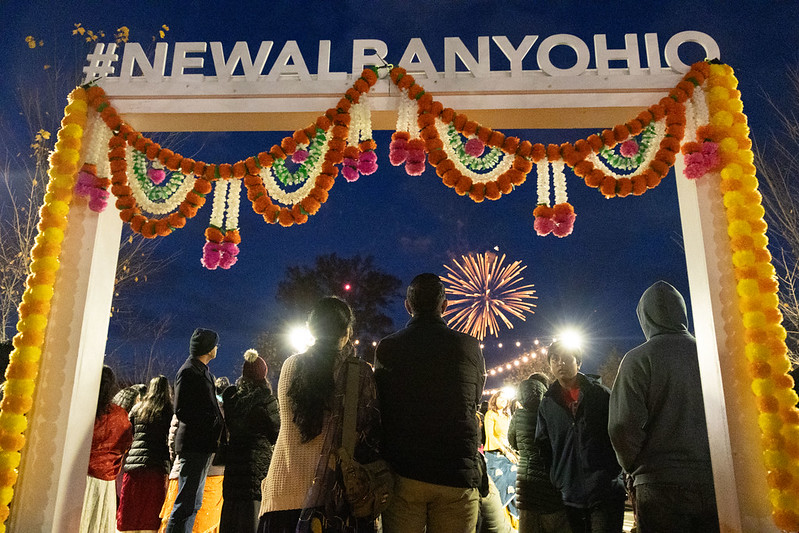 People gathered under a decorated arch with the hashtag "#NEWHEREOHIO," watching fireworks in the evening sky.