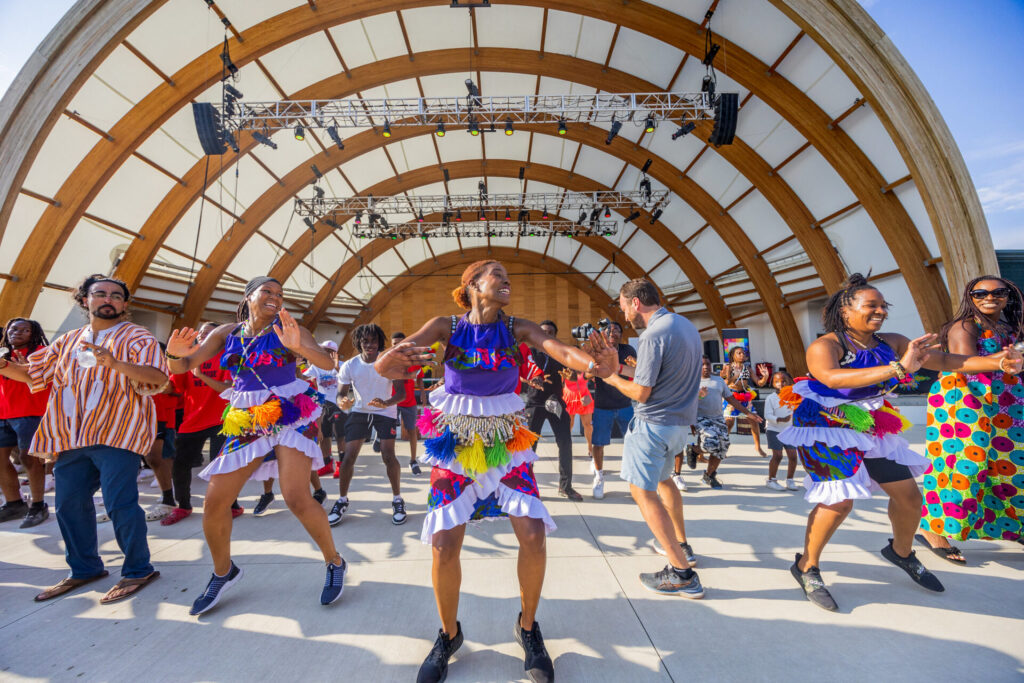 A group of people enjoying a vibrant dance performance under a large open-air stage with a curved wooden canopy.