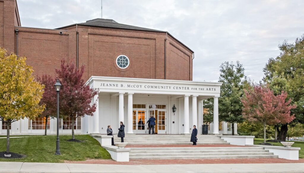 A brick building with the entrance to the Jeanne B. McCoy Community Center for the Arts, featuring white columns and steps, surrounded by trees with autumn foliage.