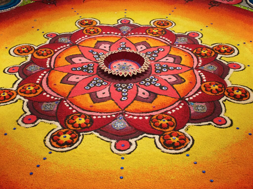 Colorful rangoli design with intricate patterns in red, orange, and yellow hues, featuring circular and floral motifs.