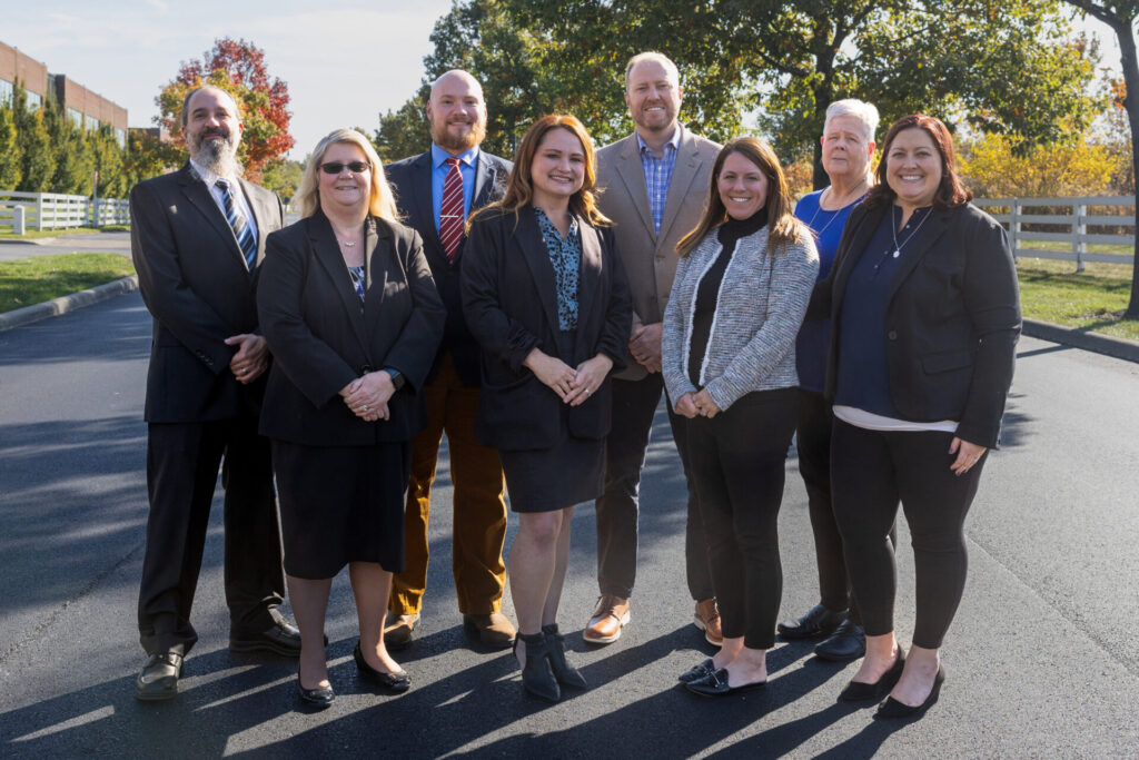 A group of eight people standing outdoors on a sunny day, all smiling and dressed in business attire.