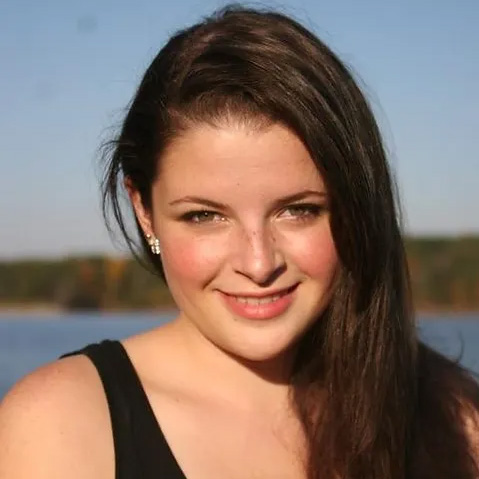 A woman with long brown hair smiling in front of a blurred outdoor background.