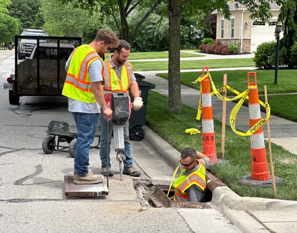 Construction workers in high-visibility vests working on a street curb near traffic cones and caution tape.