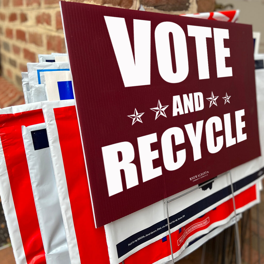 A stack of campaign signs with the top sign displaying "Vote and Recycle" in bold letters.