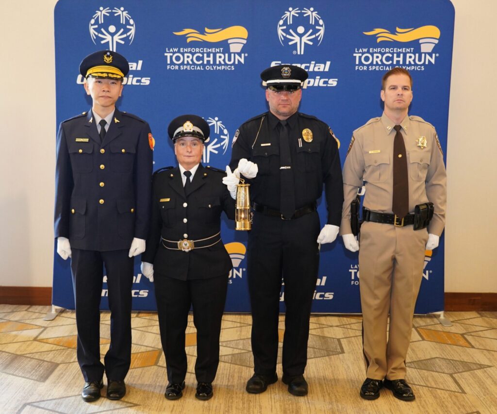 Four uniformed law enforcement officers standing in front of a Special Olympics Law Enforcement Torch Run banner, one holding a ceremonial lantern.