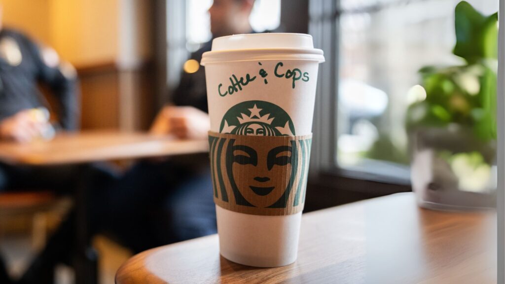 A Starbucks cup on a table with the words "Coffee & Cops" written on it; blurred background with people sitting.