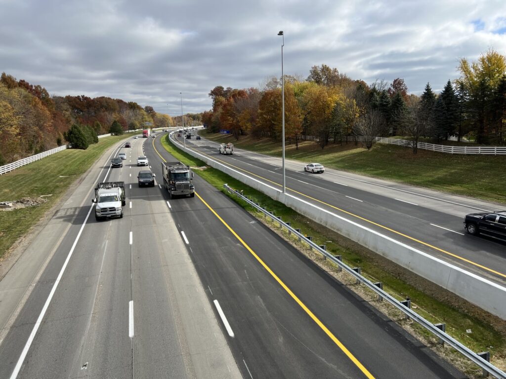 A multi-lane highway with cars and trucks driving, surrounded by a grassy landscape and autumn trees under a cloudy sky.