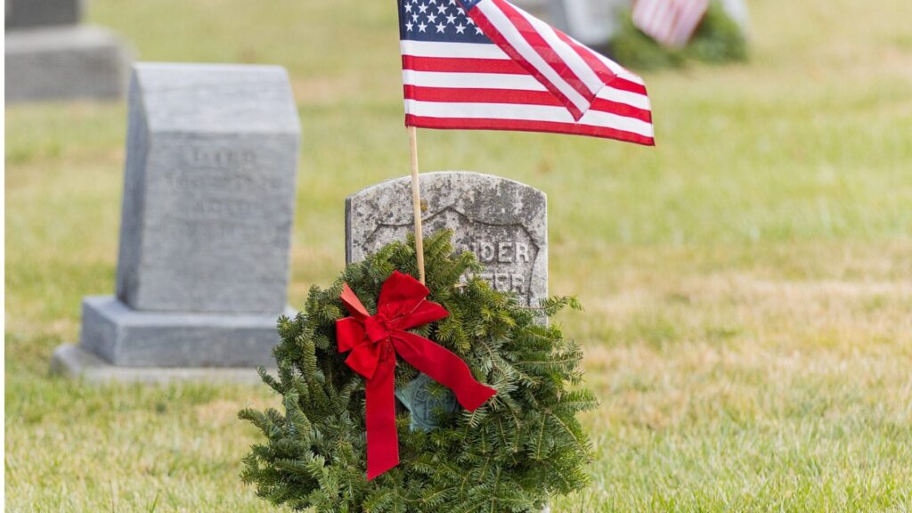 Tombstone with a Christmas wreath and an American flag on a cemetery lawn.