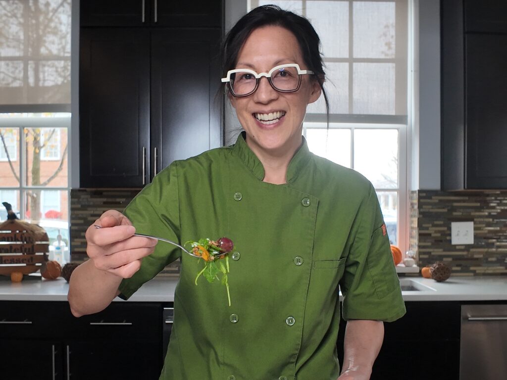 A smiling person in a kitchen wearing a green chef's coat, holding a fork with salad.