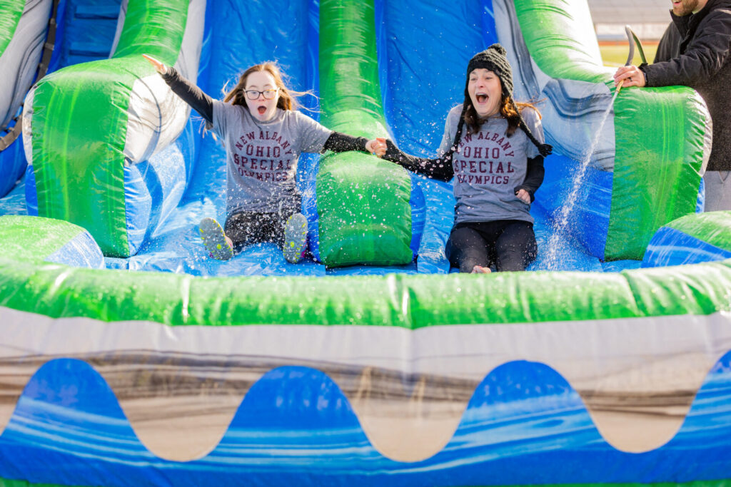 Two people sliding down an inflatable water slide, wearing "New Albany Ohio Special Olympics" shirts, with expressions of excitement.