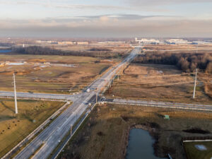 Aerial view of a rural intersection with surrounding fields, power lines, and distant industrial buildings under a partly cloudy sky.