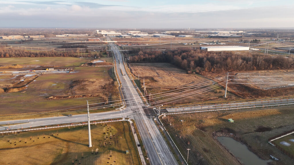 Aerial view of a rural intersection with surrounding fields, power lines, and industrial buildings in the distance.